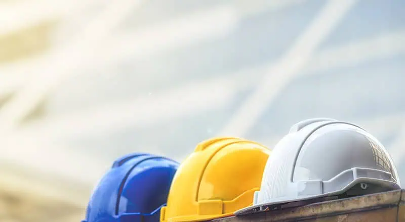 A blue, a yellow, and a white hardhat sitting beside each other on the ledge of a steel beam in front of a building in daylight.