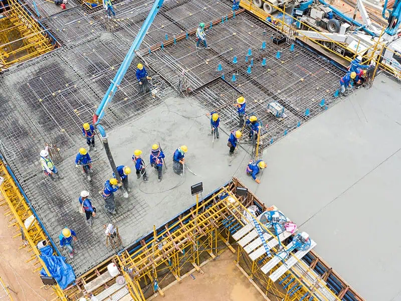 Vue aérienne d'un chantier de construction où des ouvriers en uniformes bleus et casques de protection jaunes utilisent une pompe à béton.